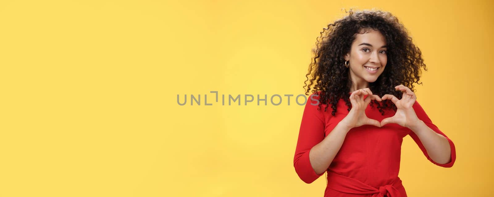 Waist-up shot of romantic and cute pretty girlfriend with curly hair in red dress showing heart sign over chest and smiling broadly confessing in admiration and love posing over yellow background by Benzoix
