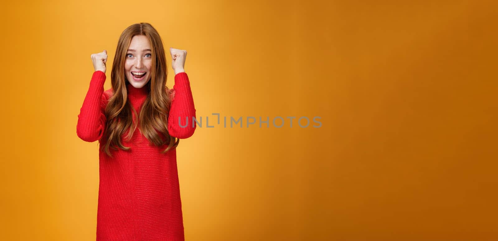Happy delighted and cheerful ginger girl with freckles and blue eyes triumphing yelling yes with broad smile clenching raised fists in joy and celebration of success against orange background. Copy space