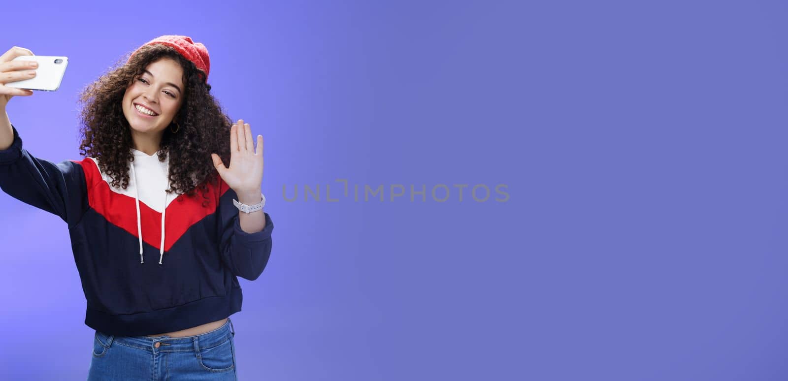 Studio shto of stylish cute female vlogger with curly hair in winter red hat smiling and waving hello at smartphone camera as recording video, making interesting content to post online over blue wall.