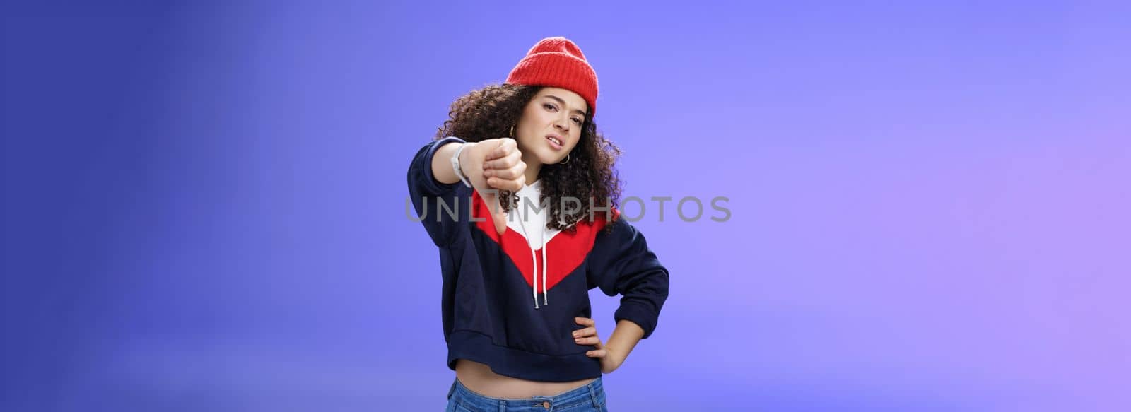 Nah dislike. Portrait of annoyed and unimpressed arrogant cool girl with curly hair tilting head with distain and scorn showing thumbs down in disappointment grimacing, posing over blue wall by Benzoix