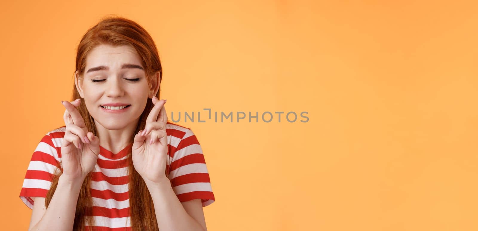 Close-up dreamy cute tender ginger girl make wish, cross fingers good luck closed eyes bite lip eager win lottery, supplicating, praying dream come true, stand orange background believe miracle.