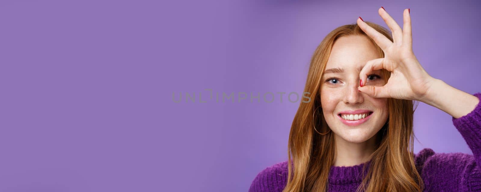 Headshot of optimistic and playful kind cute redhead female with freckles and white perfect smile showing okay or zero sign on eye, grinning as peeking through hole at camera over purple background.