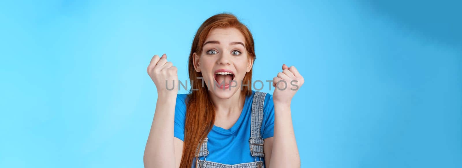 Close-up cheerful lucky redhead woman win lottery, fist pump yell yeah hooray, winning bet, smiling gladly rejoicing excellent news, celebrating success, triumphing happily, stand blue background by Benzoix