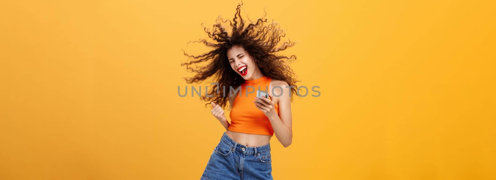 Portrait of joiyful carefree and happy charming european woman with curly hairstyle laughing out loud holding smartphone wearing wireless earbuds listening awesome music via app over orange wall. Technology concept