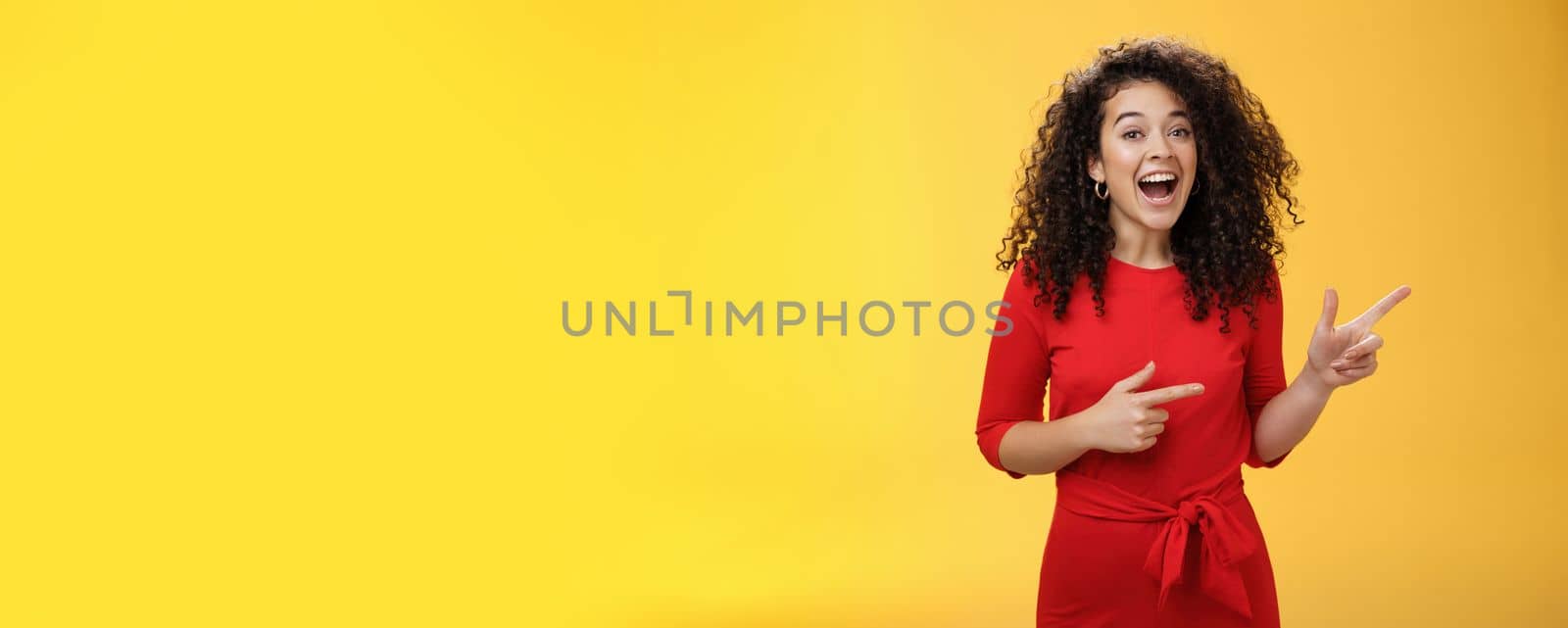 Thrilled and happy joyful young charismatic woman with curly hair in red dress laughing out loud and smiling amazed, delighted pointing at upper right corner impressed over yellow background.