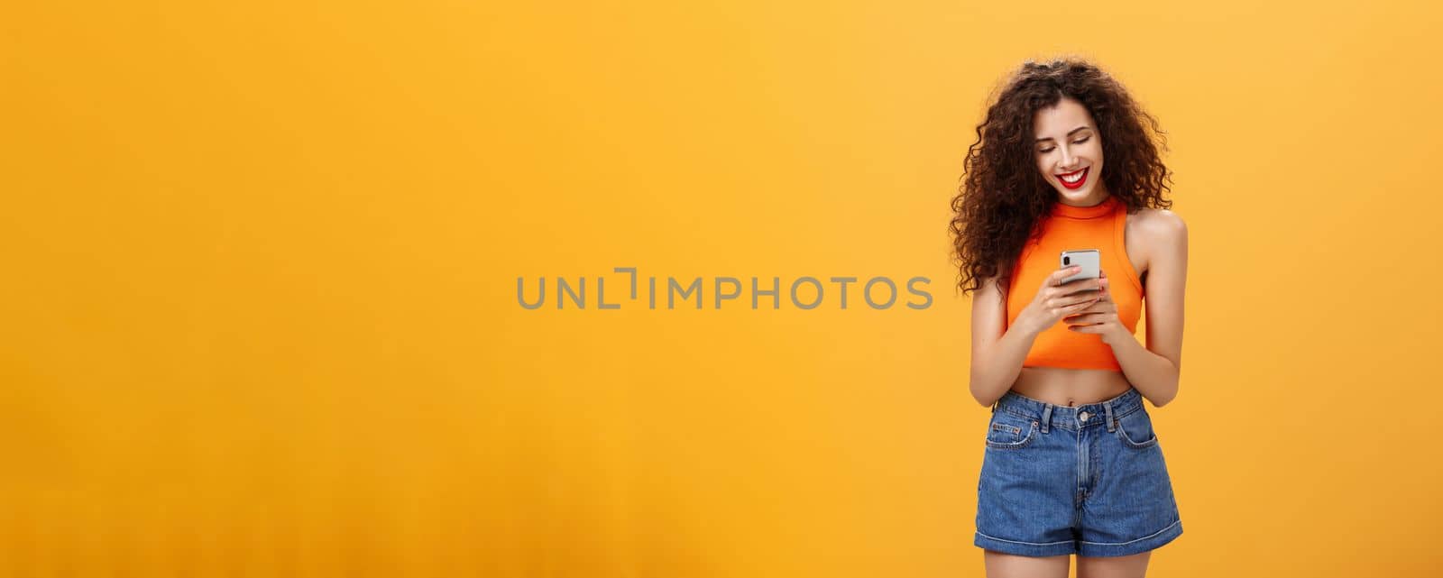 Girl spending time in internet texting friend messages via smartphone laughing while looking at device screen standing happy and upbeat over orange background in cropped top and denim shorts by Benzoix