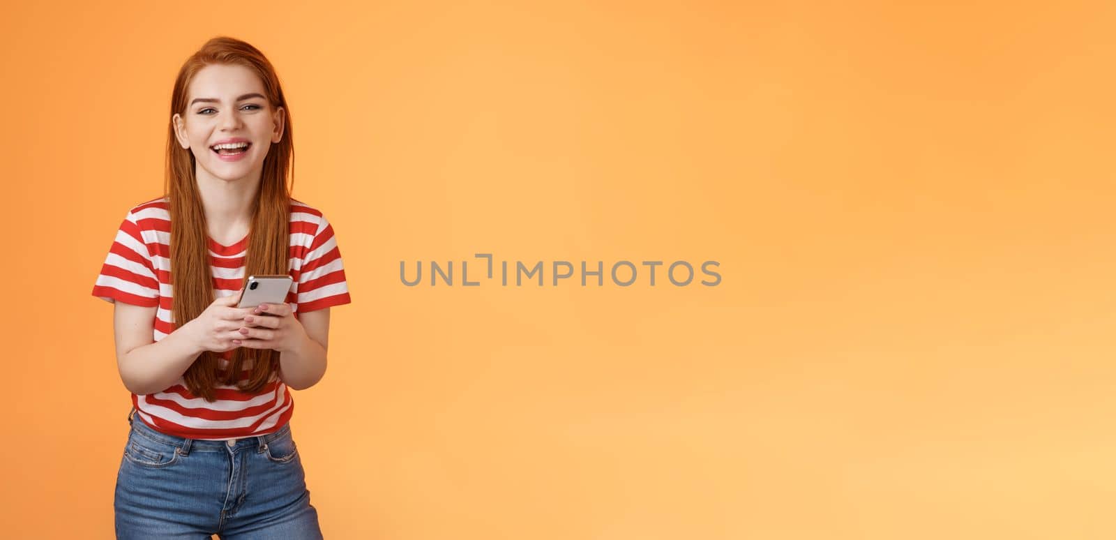 Carefree attractive woman red long hair laughing amused, having fun, chatting friend, hold smartphone, look camera sincere rejoice smile, texting joke, found funny meme internet, orange background.
