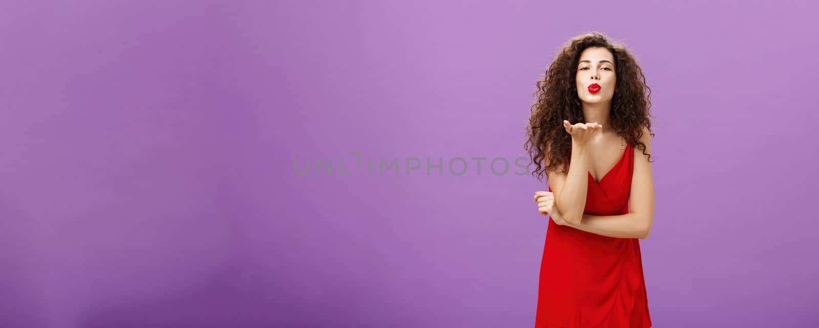 Woman sending passionate kiss being hopeless romantic. standing in elegant red dress with curly hairstyle and makeup blowing mwah with smile and folded lips holding palm near mouth over purple wall.