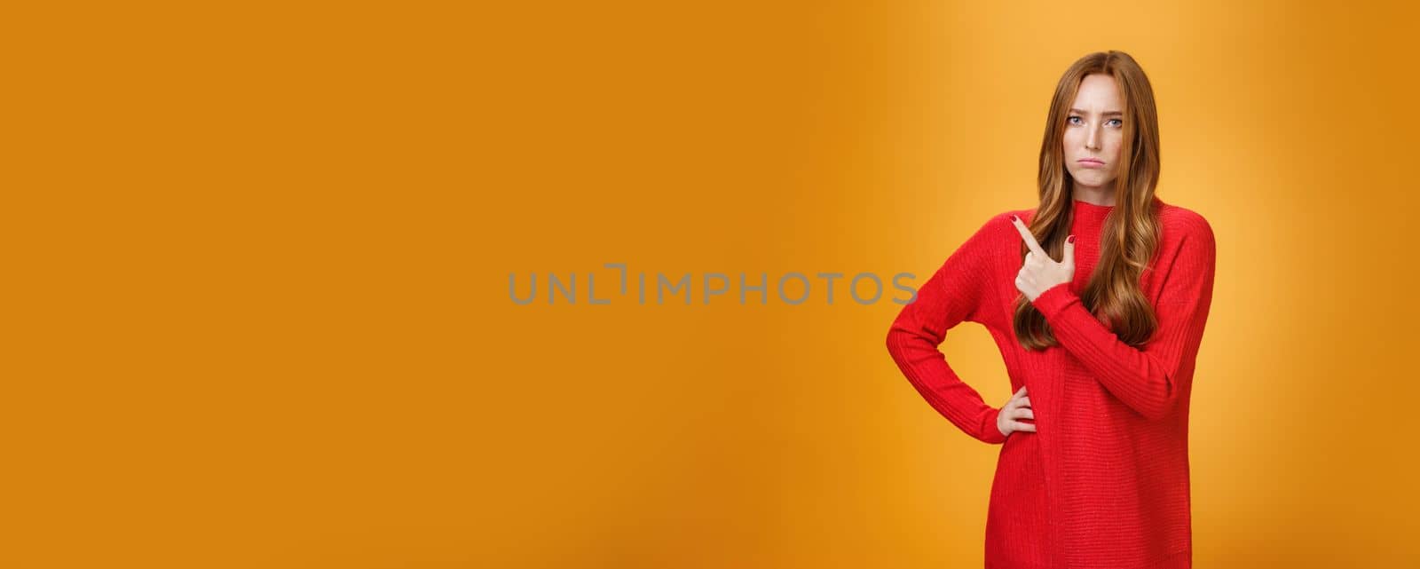Upset and disappointed young ginger girl in red sweater pointing at upper left corner or behind frowning looking gloomy and angry, standing jealous and moody against orange background.