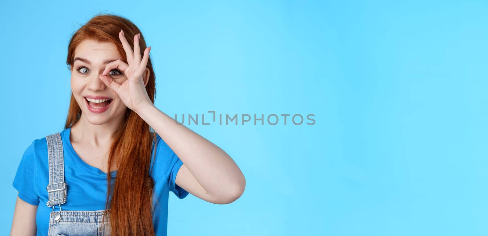 Cheerful upbeat redhead european female feel lucky inspired, look through circle okay on eye, smiling broadly amused, look wondered admire cool awesome sales, stand blue background. Copy space