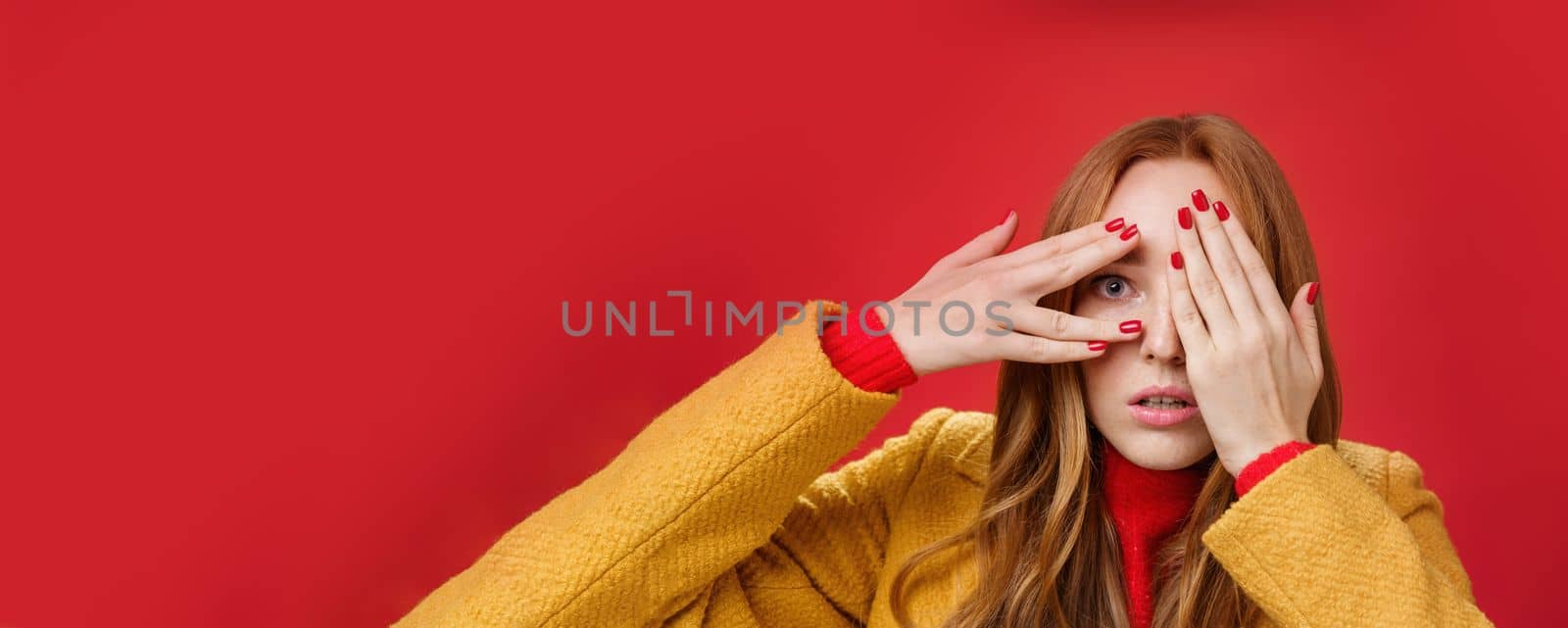 Lifestyle. Stunned and shocked redhead woman cannot believe what terrible thing she sees covering face with palms open mouth from shook and peeking through fingers anxious and scared over red background.