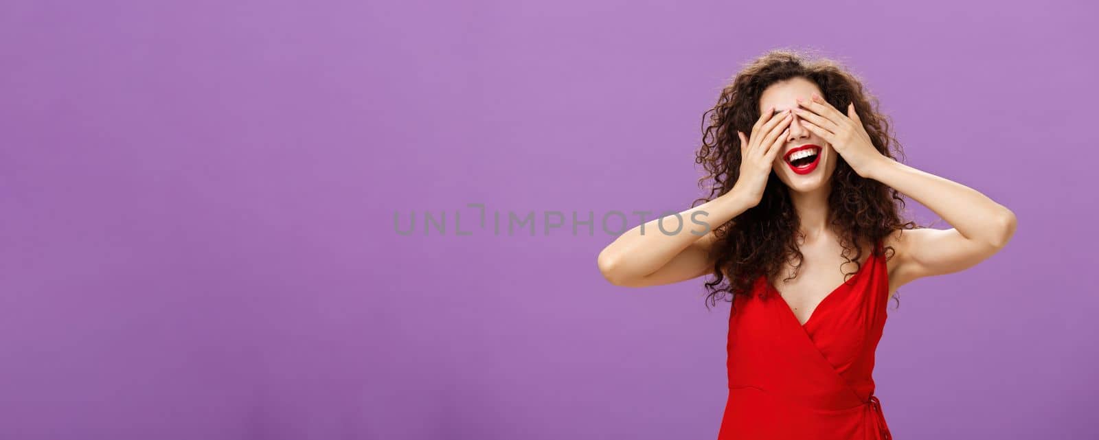 Wife waiting for husband present gift for anniversary closing eyes with palms and smiling broadly peeking through fingers being charmed and excited standing in seductive red dress over purple wall. Emotions concept