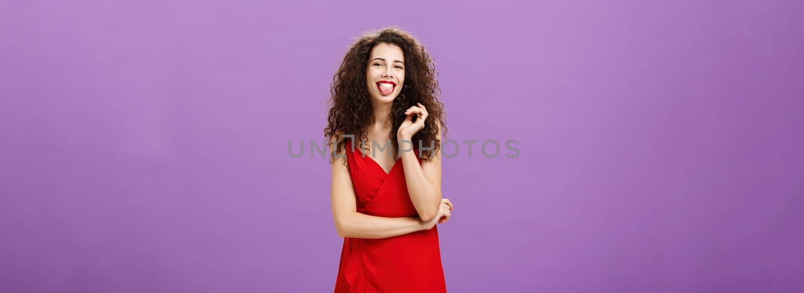 Studio shot of playful and joyful artistic caucasian female. student in red evening dress sticking out tongue happily having fun playing with curl standing over purple background careless and carefree.