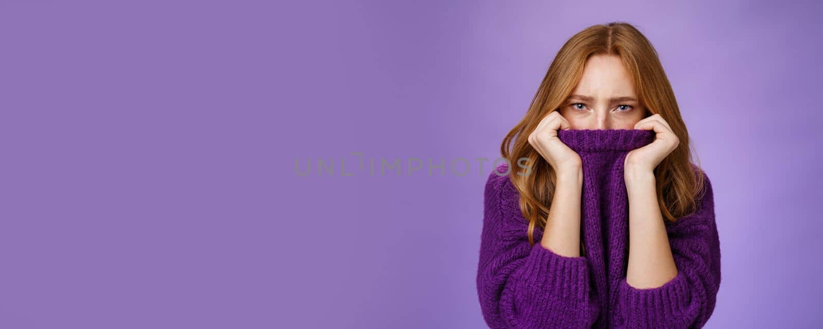 Gloomy upset redhead girlfriend pulling collar of purple sweater on nose, frowning and squinting offended, being aggrieved and resent, sulking moody and displeased over violet background.