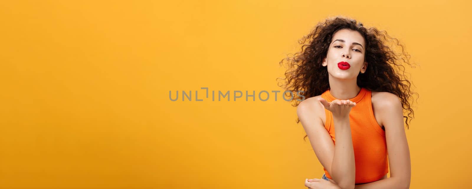 Flirty confident and sensual stylish woman with curly hairstyle and red lipstick folding lips holding palm near mouth sending kiss while hair flicks on air being in romantic mood over orange wall by Benzoix