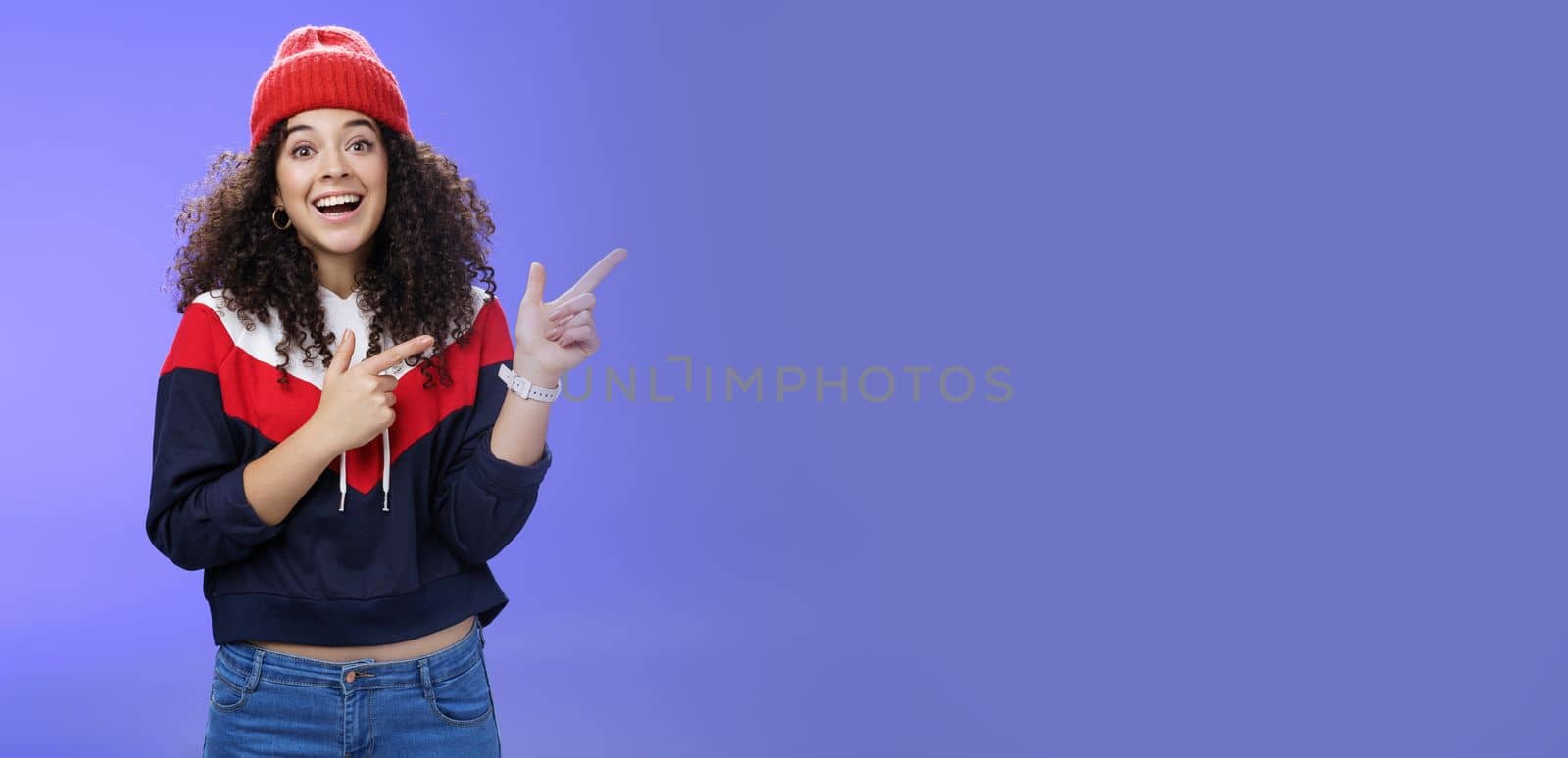 Lifestyle. Enthusiastic friendly-looking attractive young 20s woman with curly hair in warm beanie and sweatshirt smiling open mouth amazed and delighted as pointing at upper right corner over blue background.