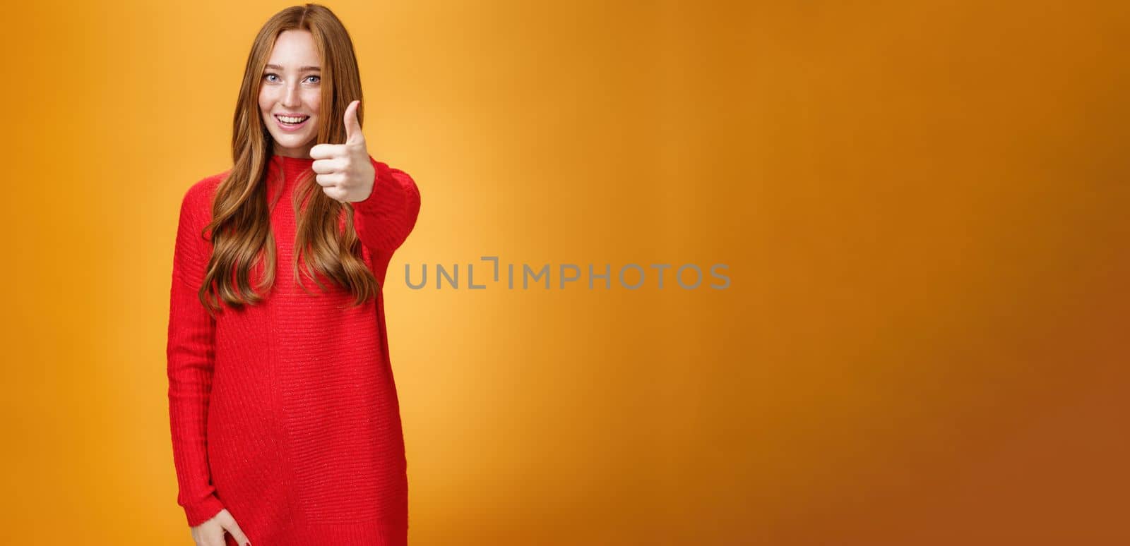 Nice job good. Portrait of delighted and energized confident ginger woman in red sweater pulling thumb up at camera and smiling, liking idea, approving and accepting it over orange background. Copy space