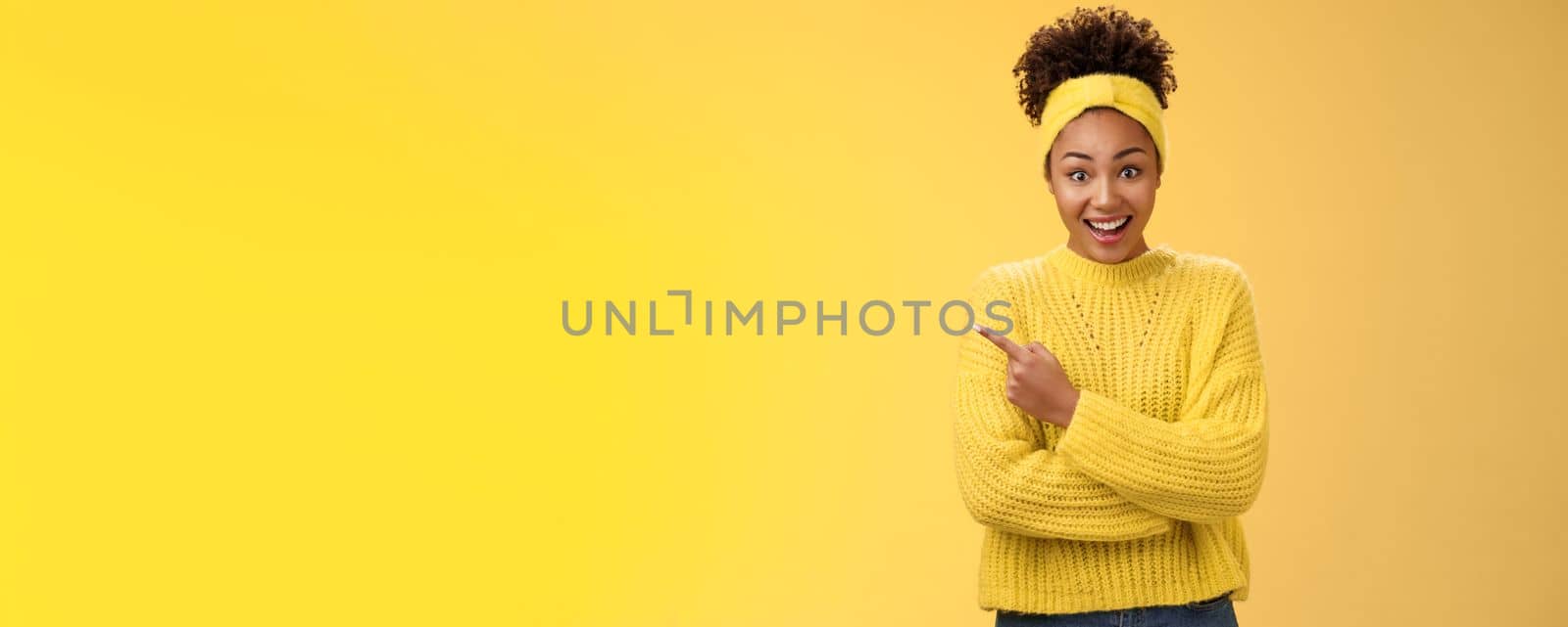 Amused charismatic smiling black cute girl. in headband sweater widen eyes drop jaw astonished hear about awesome interesting new place talking friend standing yellow background pointing left amazed.