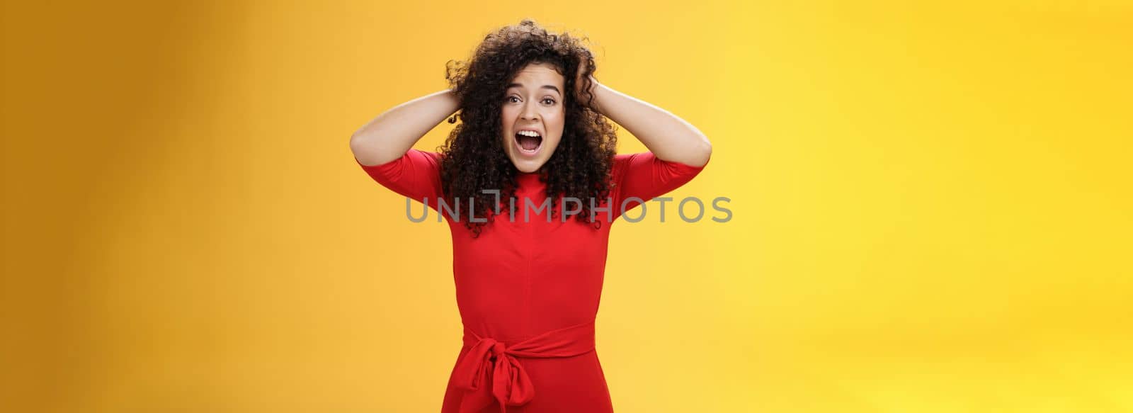 Girl feeling pressure standing anxious in panic holding hands on curly hair yelling at camera disturbed, freaked out being tensed and upset with bad situation, standing troubled over yellow background.