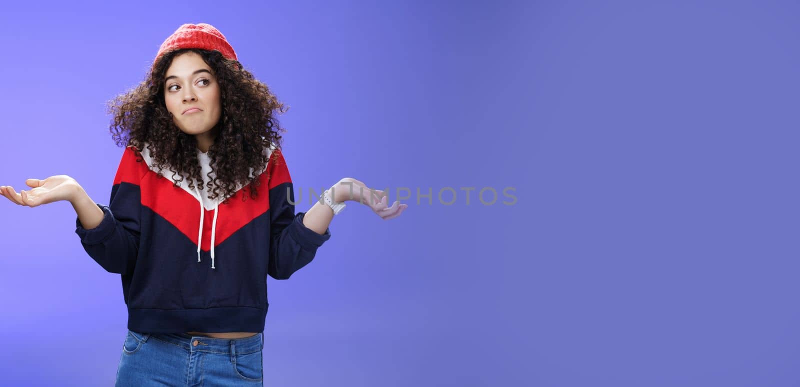 Not getting clue. Cute silly woman with curly hair shrugging with pursed lips and unbothered expression holding hands sideways looking right being unaware and confused, uncertain what answer by Benzoix