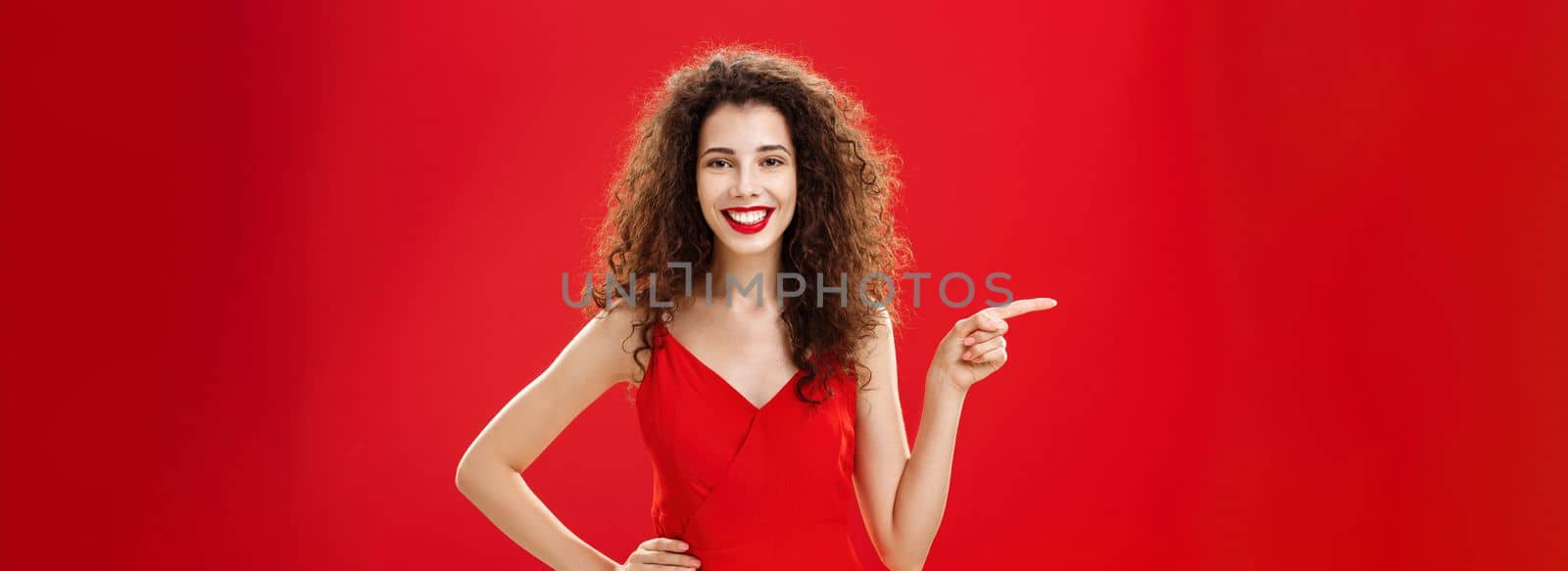 Girl suggesting ask her boyfriend. Charming elegant and stylish young female with curly hairstyle in red evening dress holding hand on hip and pointing left while showing friend where buy clothes.