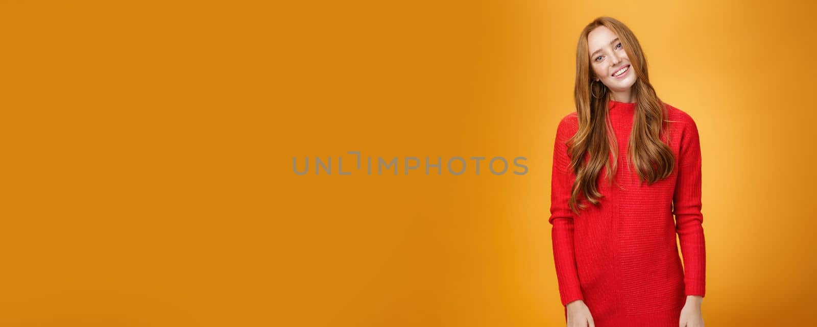 Lifestyle. Attrative and stylish elegant busiensswoman with ginger hair in red knitted dress tilting head and smiling flirty and happily posing over orange background as listening with interest to conversation.
