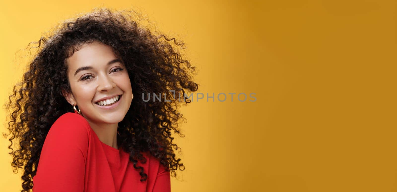 Romantic and flirty young cute caucasian girlfriend with curly hair in red blouse making silly pose smiling joyfully and lifting shoulder coquettish smiling broadly at camera over yellow wall. Love, relationship, seduction concept