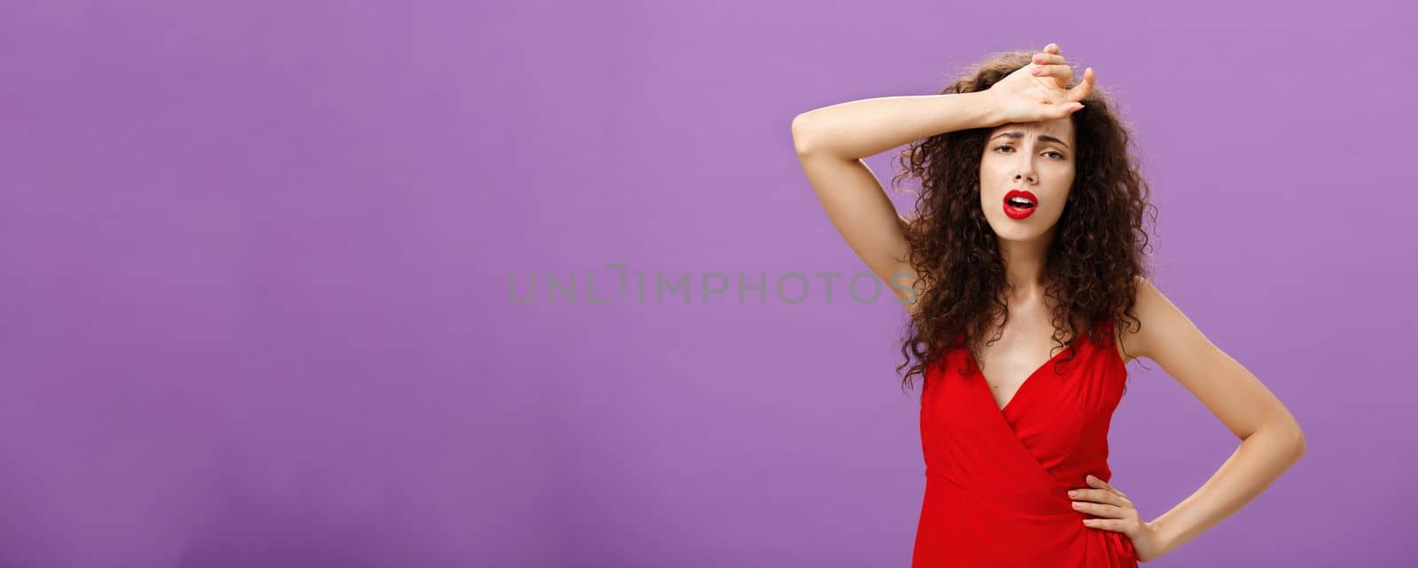 Woman fainting feeling bad whiping sweat of forehead standing drained and exhausted over purple background in red stylish dress expressing gloomy and unhappy feelings wanting some help. Copy space