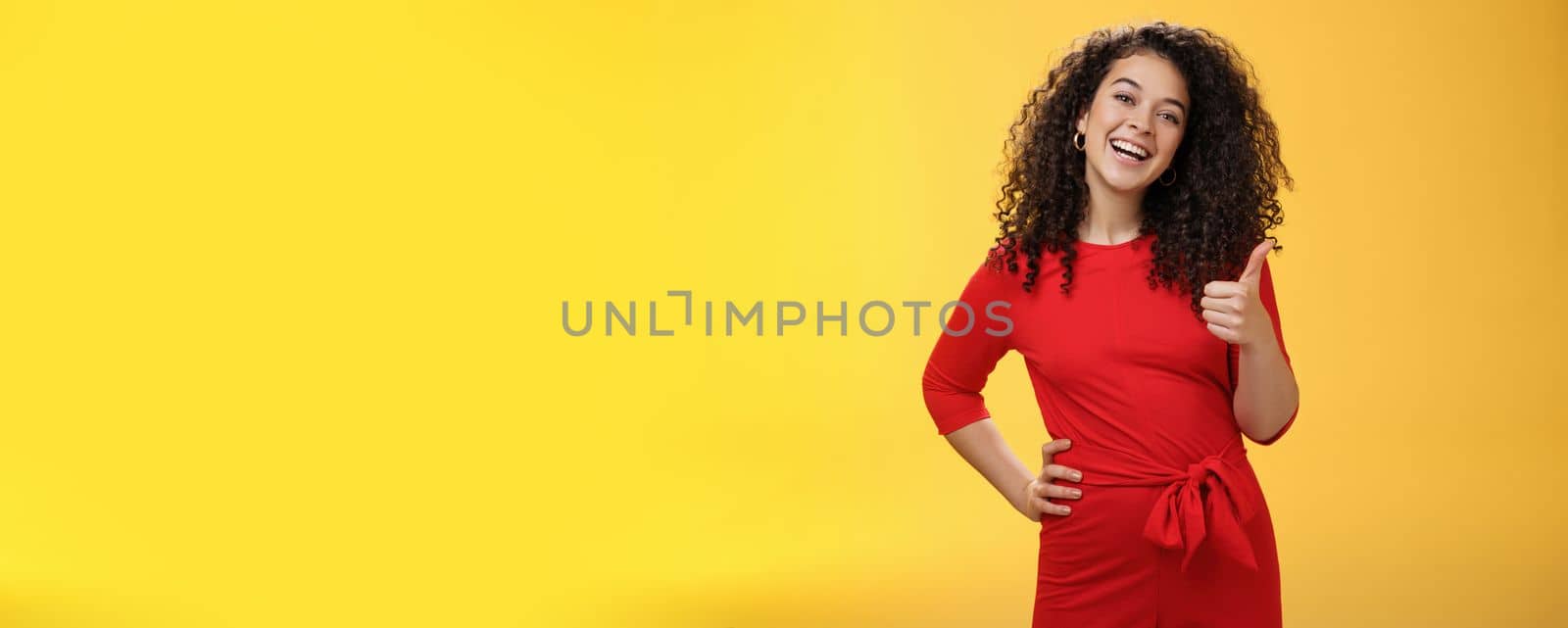 Portrait of happy optimistic young curly-haired woman in red dress laughing joyfully, showing thumbs up in approval and like gesture, delighted with awesome idea, accepting plan over yellow wall by Benzoix