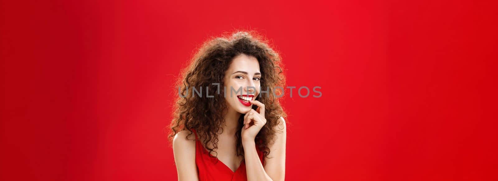 Waist-up shot of sexy and sensual adult girlfriend. with curly hair in elegant dress biting finger and smiling flirty seducing hot guy during formal event posing against red background romantically.