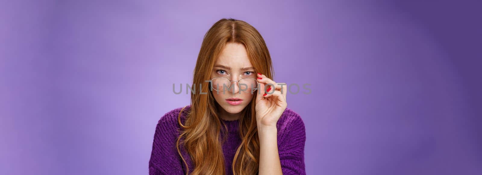 Questioned and unsure cute smart female detective with ginger hair looking suspicious from under forehead taking off glasses and looking at camera doubtful and uncertain over purple background by Benzoix