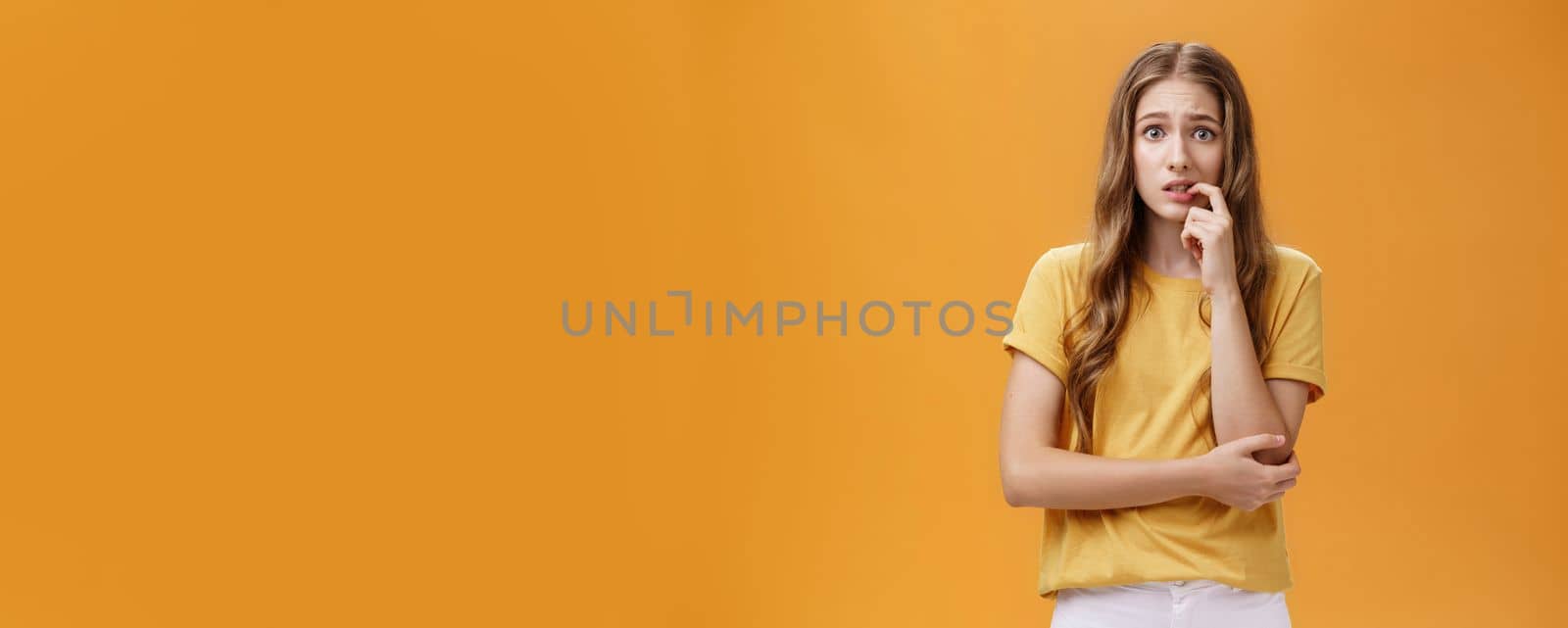 Girl worried mom react on new tattoo. Portrait of anxious and nervous silly insecure young female with wavy natural hairstyle biting finger panicking looking concerned and troubled over orange wall by Benzoix