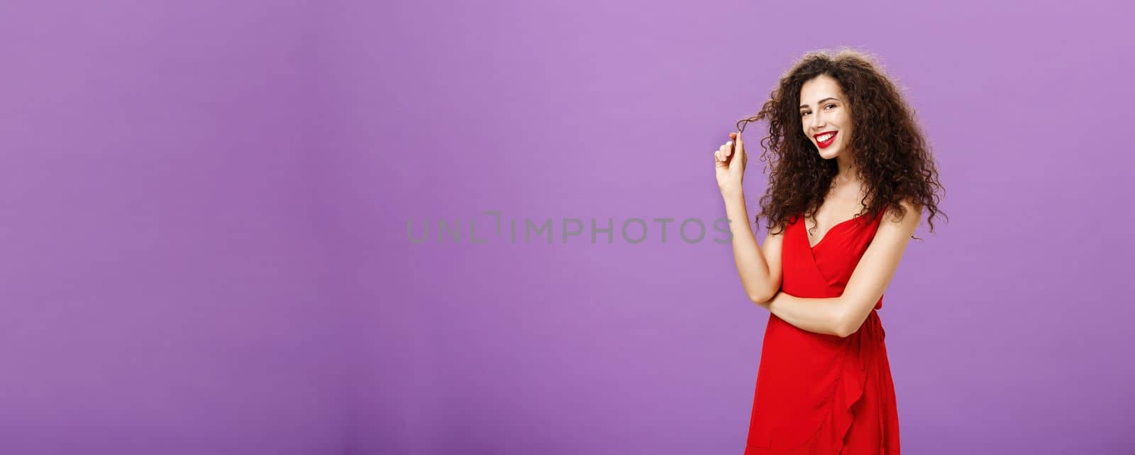 I have my eye on you. Portrait of sensual and flirty attractive curly-haired hot female in red evening dress playing with curly and smiling at camera using seduction skill to impress handsome man over purple wall.