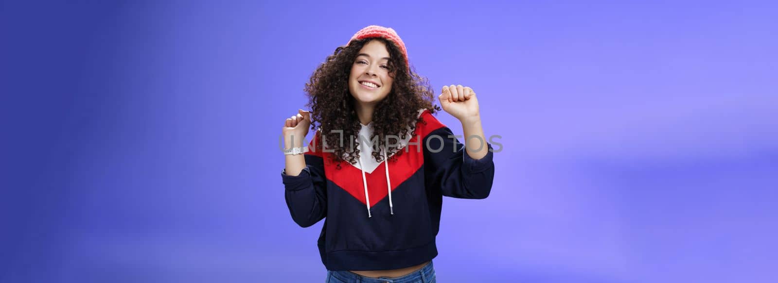 Charming carefree young 20s girl with curly haircut in warm beanie dancing and having fun joyfully over blue wall raising hands relaxed, chill smiling broadly at camera liking perfect chilly weather.