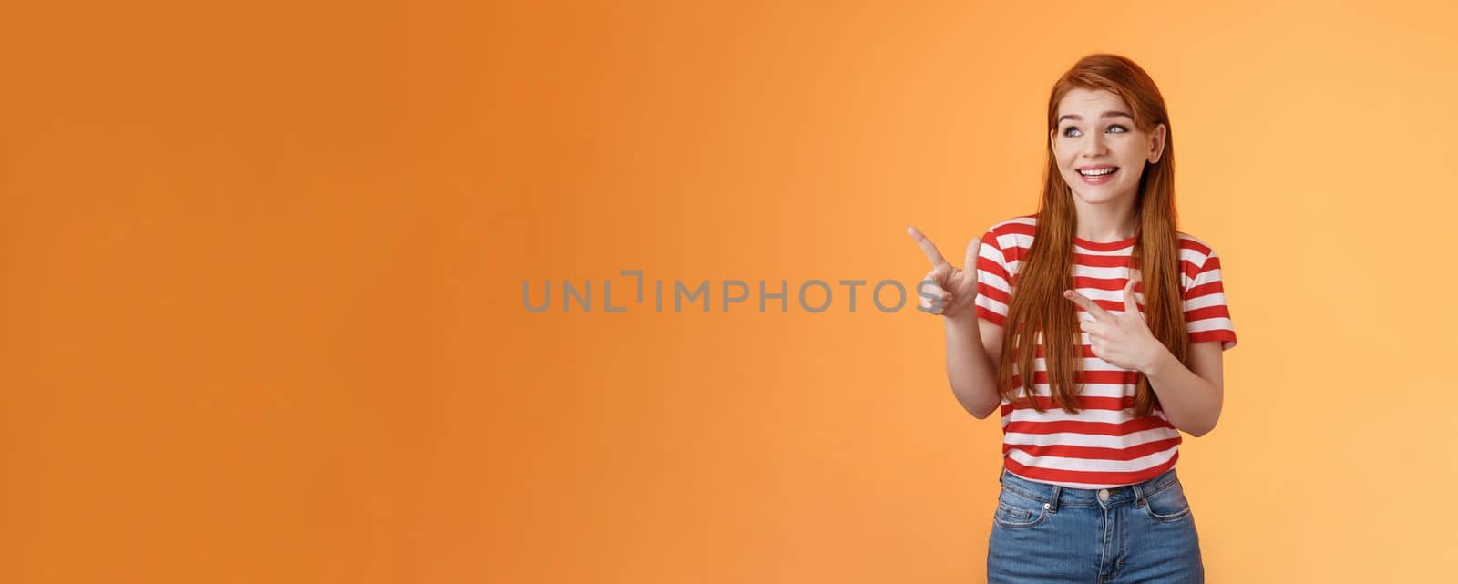 Cute attractive feminine ginger young 20s woman smiling broadly pointing look left happily, reacting amused cool new gadget feature, stand excited upbeat orange background casual urban clothes.