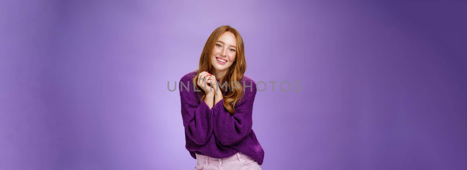 Charming and tender carefree redhead girlfriend with cute freckless holding hands together near shoulder as posing flirty and feminine smiling broadly having positive, happy attitude over purple wall.