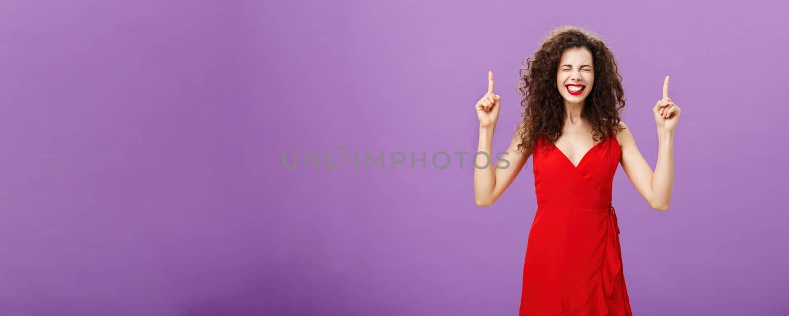 Life changing moment happening now. Joyful and happy overwhelmed good-looking elegant woman with curly hairstyle in red evening dress closing eyes grinning from happiness, pointing up over purple wall.