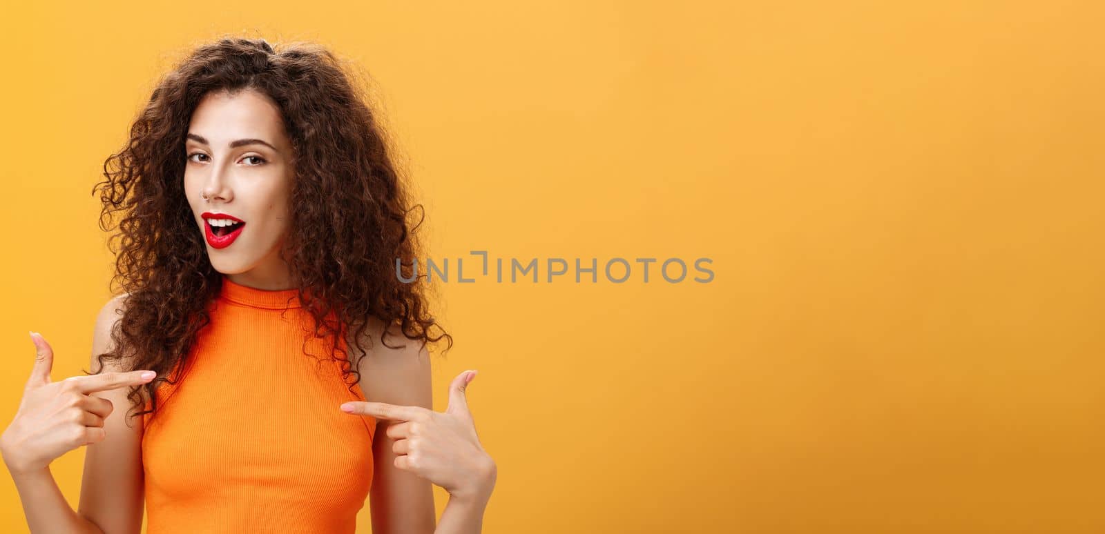 Proud and satisfied cool urban female with red lipstick. and curly hairstyle pointing at herself with self-assured expression winking bragging about skills and achievements over orange background.