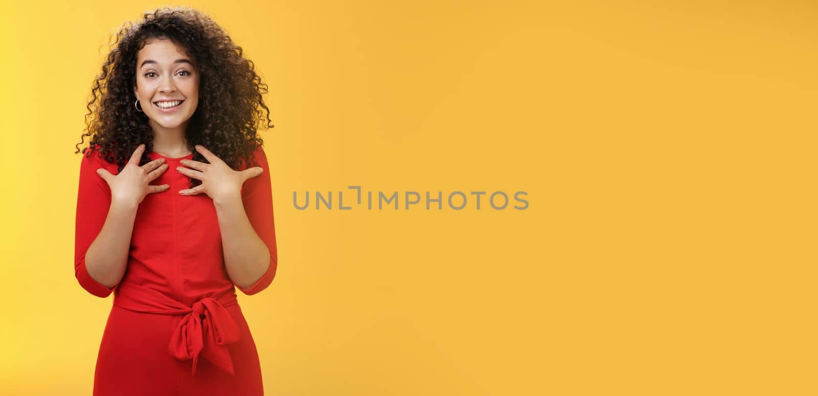 Lifestyle. Pleased and thankful tender curly-haired kind girl in red dress pressing hands to breast being surprised with unexpected tender heartwarming gift thanking smiling delighted over yellow background