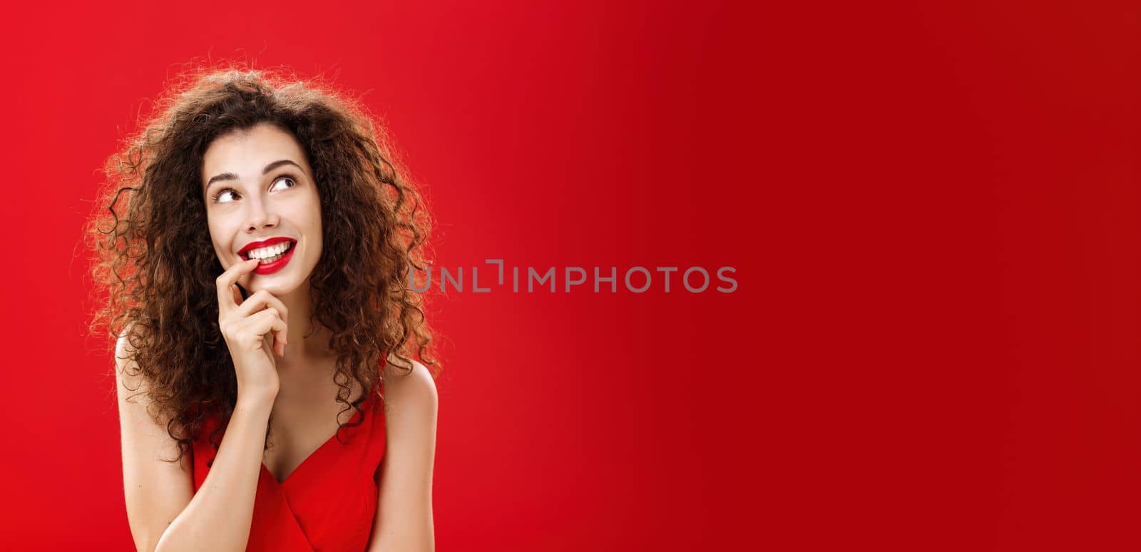 Cute charismatic and tender dreamy woman with curly hairstyle in red dress biting finger and smiling with desire in eyes looking at upper right corner daydreaming picturing object she wants by Benzoix