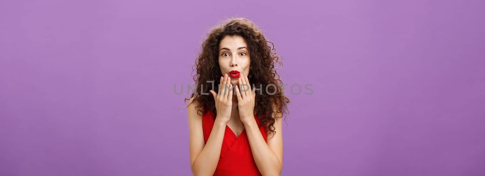 Delighted and excited luxurious adult curly-haired woman. with evening makeup in red dress folding lips in amazement holding palms on face listening carefully to interesting rumor over purple wall.