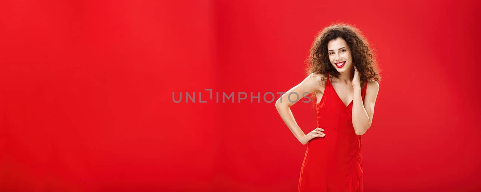 Woman posing in front of boyfriend in new stylish dress ready go out for dinner in romantic restaurant touching neck gently holding hand on hip standing in flirty and feminine pose over red background. Copy space