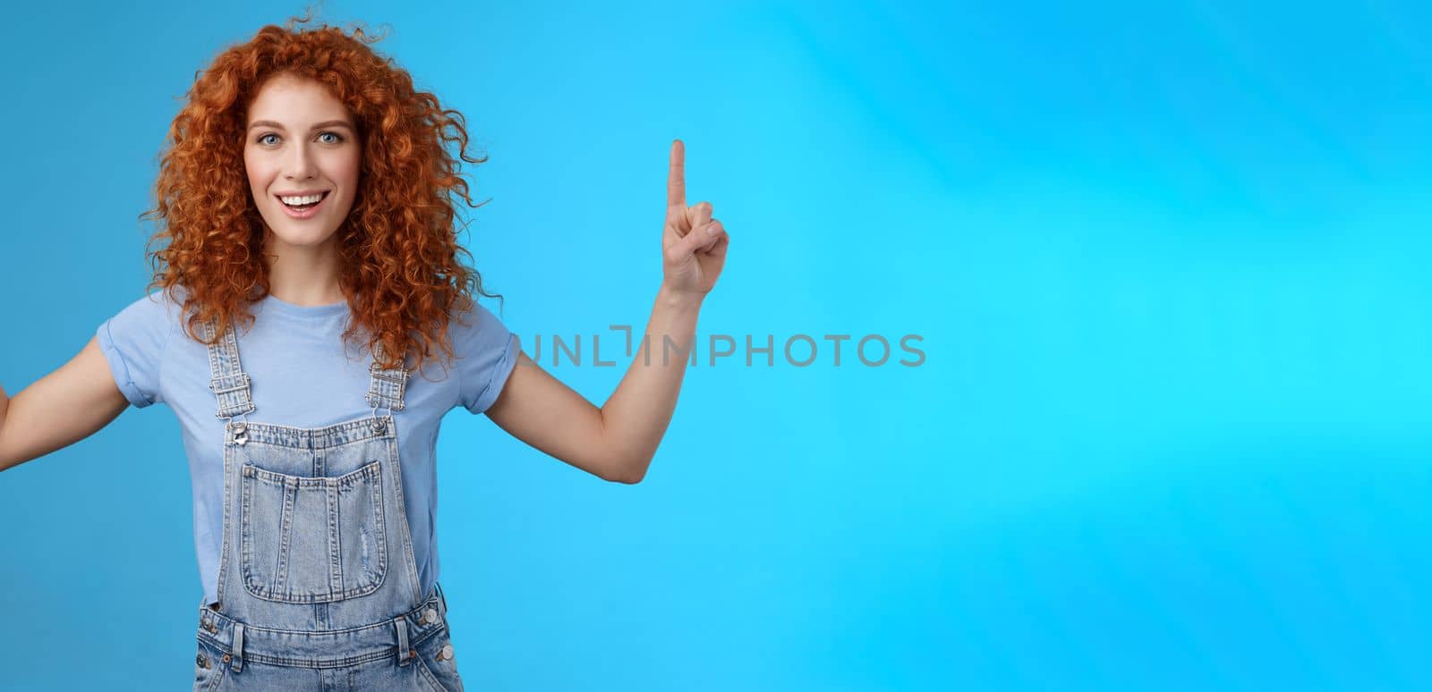 Energized enthusiastic good-looking redhead cheerful curly girl pointing up index fingers motivated present awesome copy space smiling delighted glad summer warm days near beach blue background by Benzoix