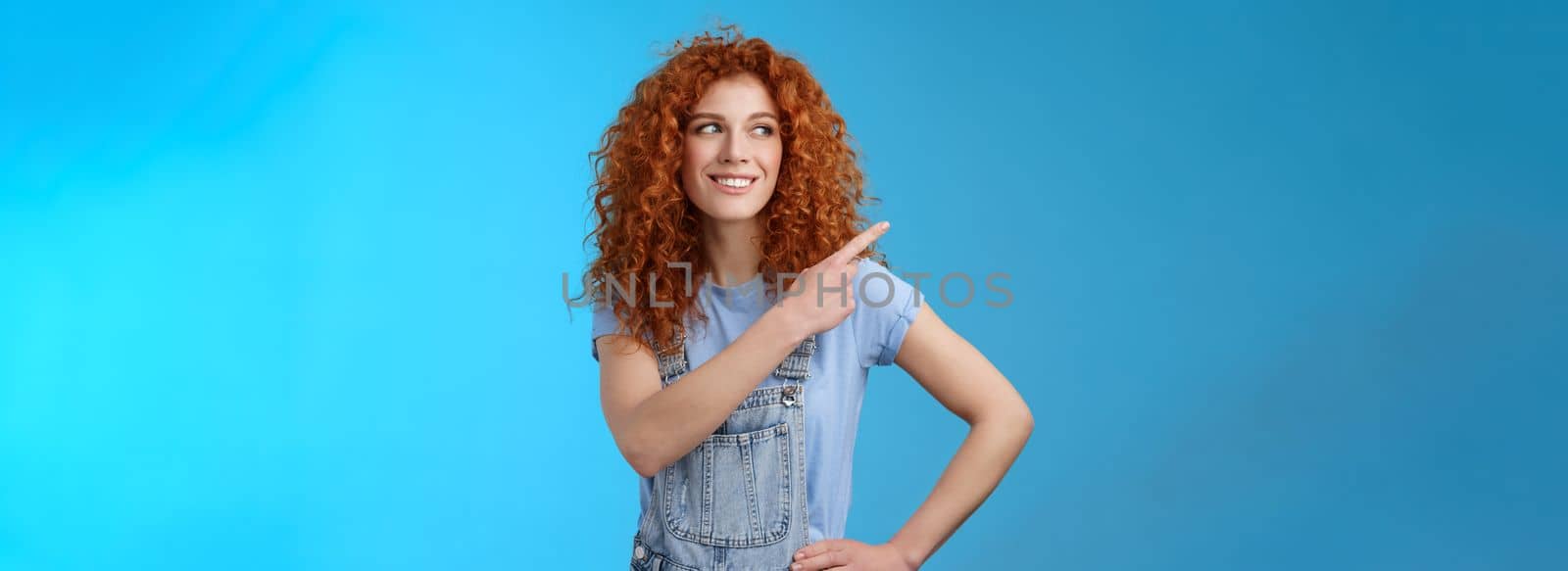 Cheerful motivated charismatic good-looking happy smiling redhead 25s curly-haired woman summer dungarees pointing upper left corner look aside pleased show promo suggest try-out. Copy space