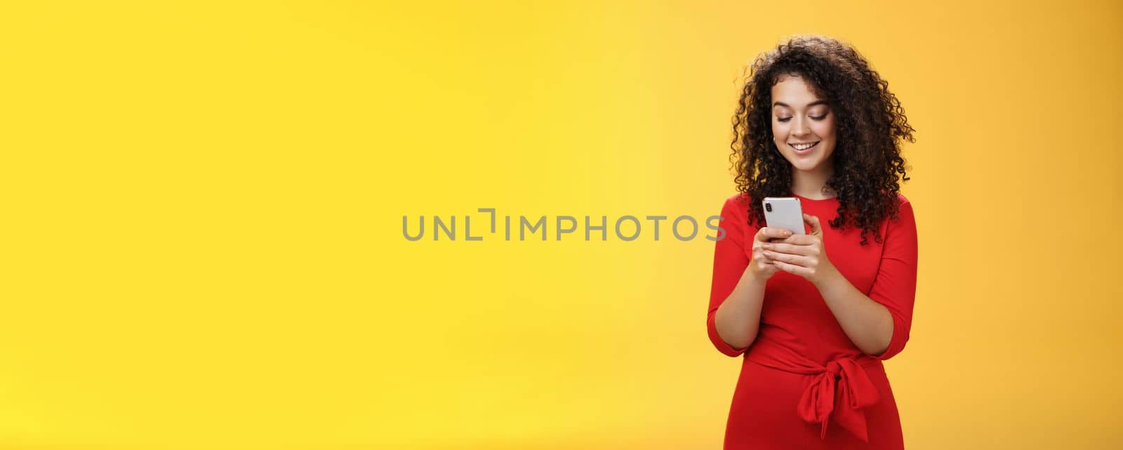 Lifestyle. Gil sending message spread news across social network having party inviting friends via smartphone holding mobile phone in hands smiling broadly at device screen as posing over yellow background.