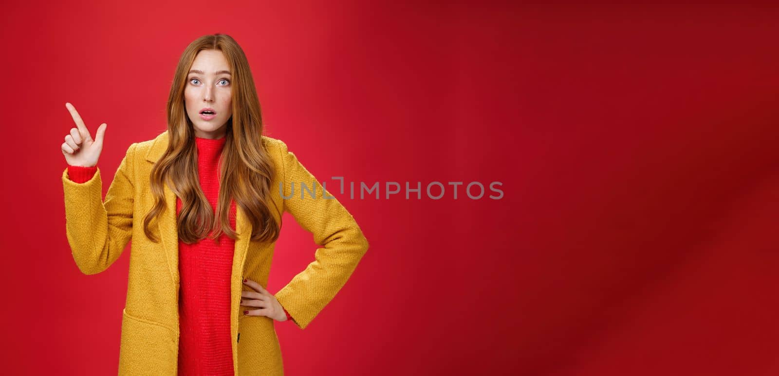 Shocked and stunned cute, confused ginger girl in yellow coat feeling worried and surprised open mouth lifting eyebrows amazed pointing at upper left corner, posing against red background.