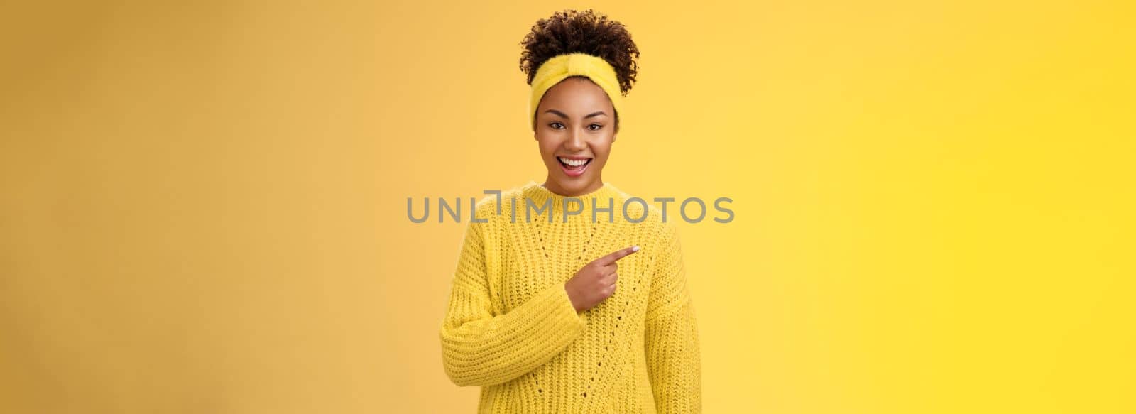 Friendly good-looking enthusiastic black charming girl in headband sweater smiling broadly amused pointing left awesome place showing you perfect blank space advertisement, posing yellow background by Benzoix