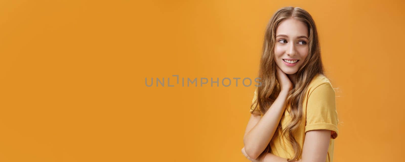 Lifestyle. Soft and tender beautiful young girl with wavy fair hair turning back and look right touching neck gently smiling silly and sincere feeling pretty and self-assured posing over orange background.