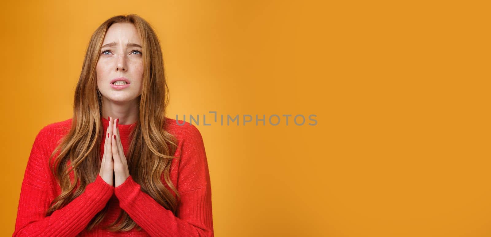 Close-up shot of anxious nervous and hopeless distressed cute redhead woman in red dress holding hands in pray looking up with supplication praying asking god help or mercy over orange wall. Copy space