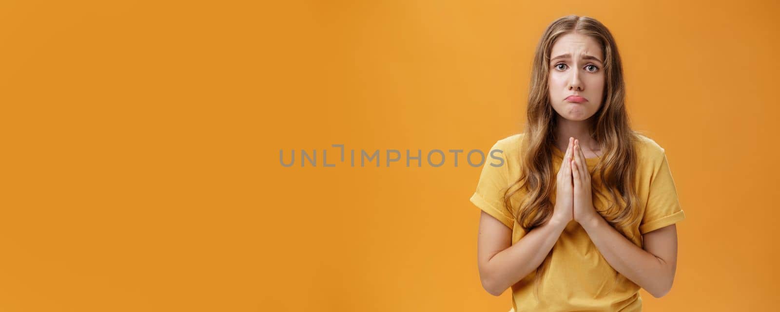 Silly girl needs help begging with hopeful puppy eyes holding hands in pray pouting and frowning feeling miserable stuck in bad situation standing upset over orange background, borrowing money.